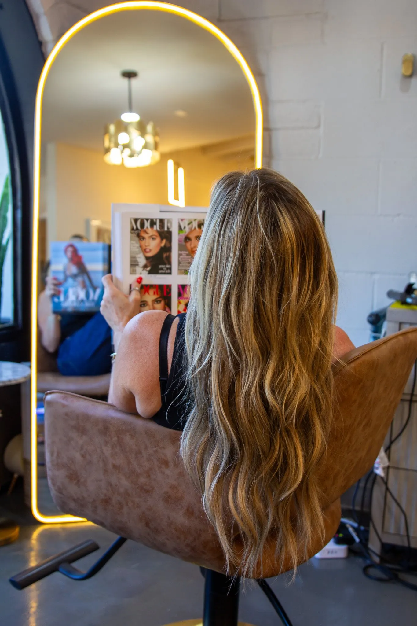 A client in the salon chair facing the lit arch mirror, long waved extensions past the back of the chair, Vogue covers on the wall behind.