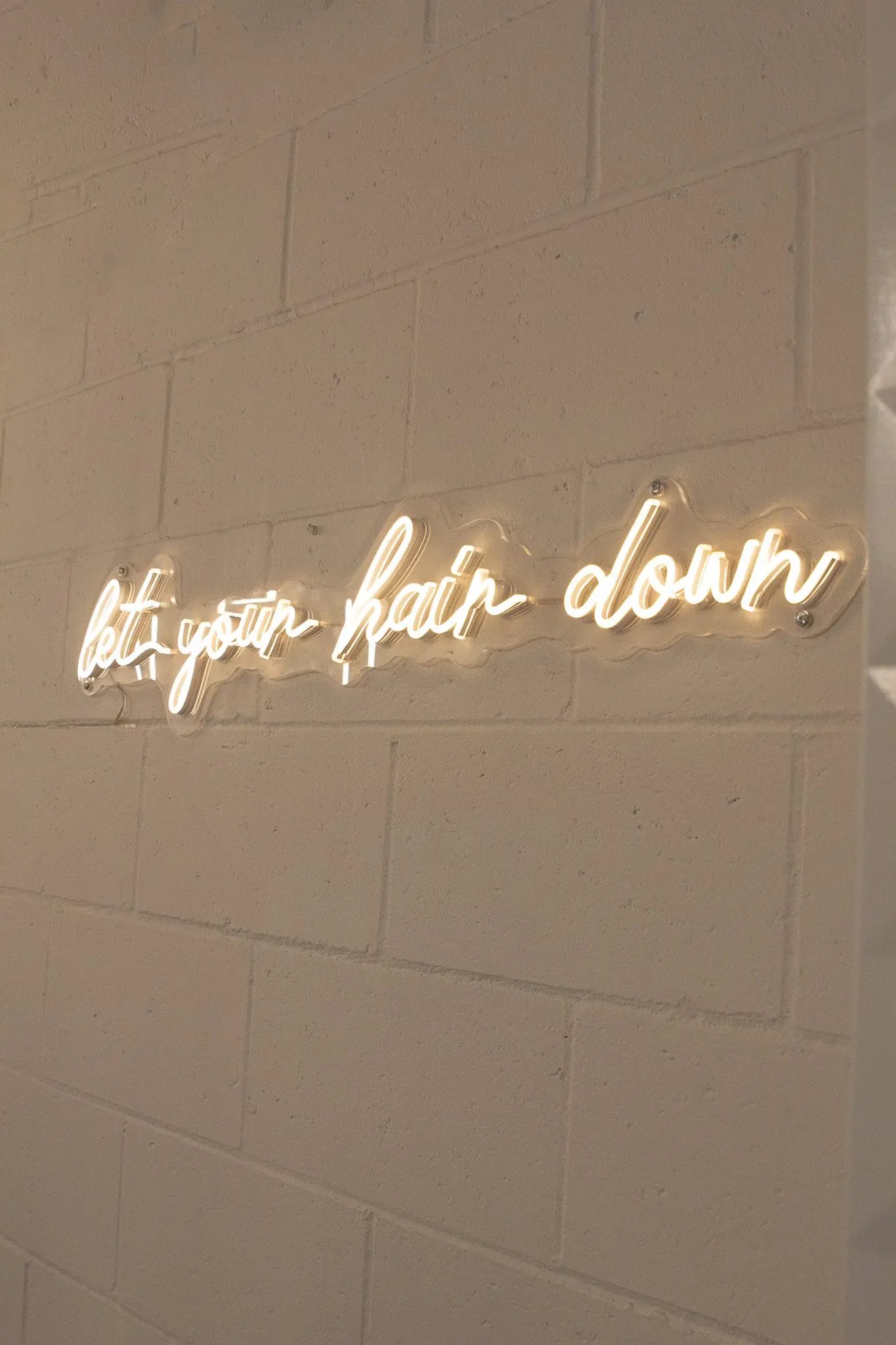 Neon script reading let your hair down on a white brick wall.